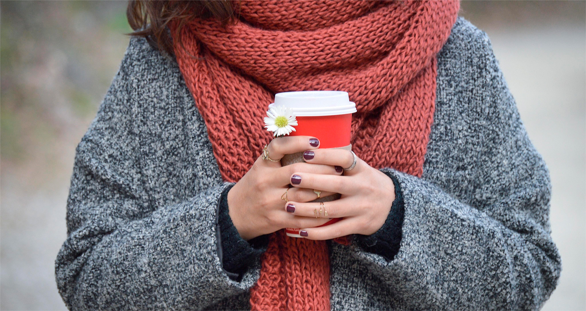 A woman in a knitted red scarf holds a cup of coffee in her hands