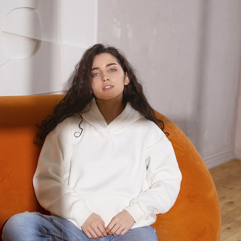 A female model in a white hoodie sits on a sofa