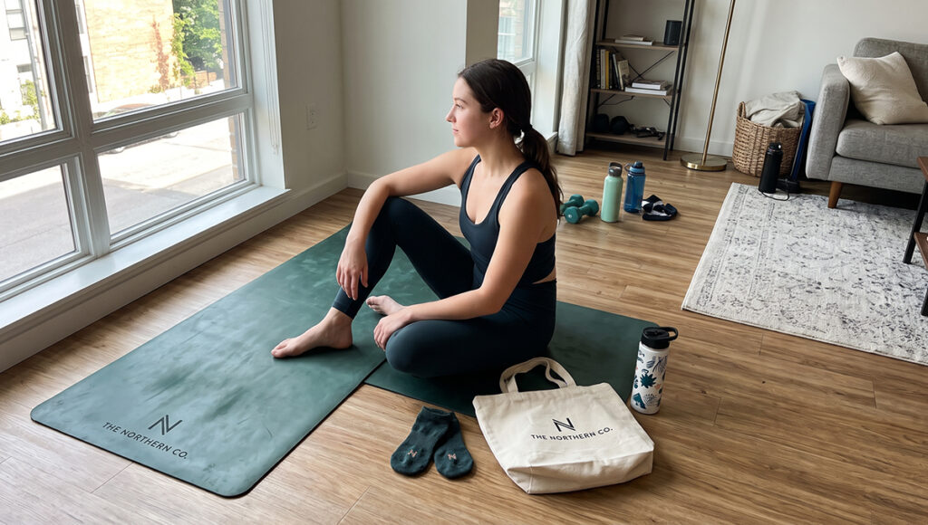 A woman in dark activewear sits on a green yoga mat in a sunlit living room, looking out the window. Arranged near her are custom branded fitness products, including the yoga mat, a pair of socks, and a canvas tote bag, all prominently featuring 